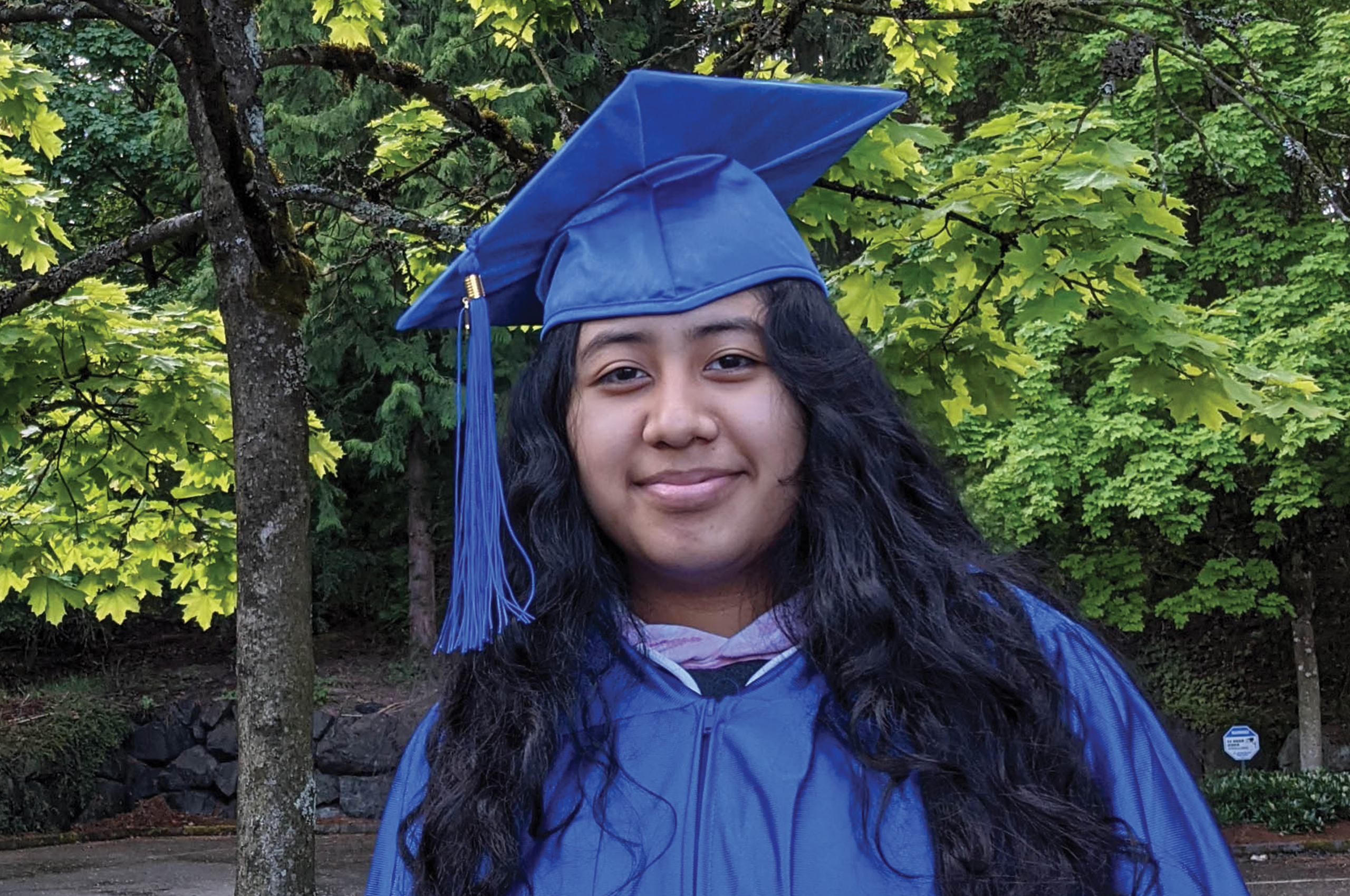 Girl in a blue cap and gown smiles in front of a tree-filled background